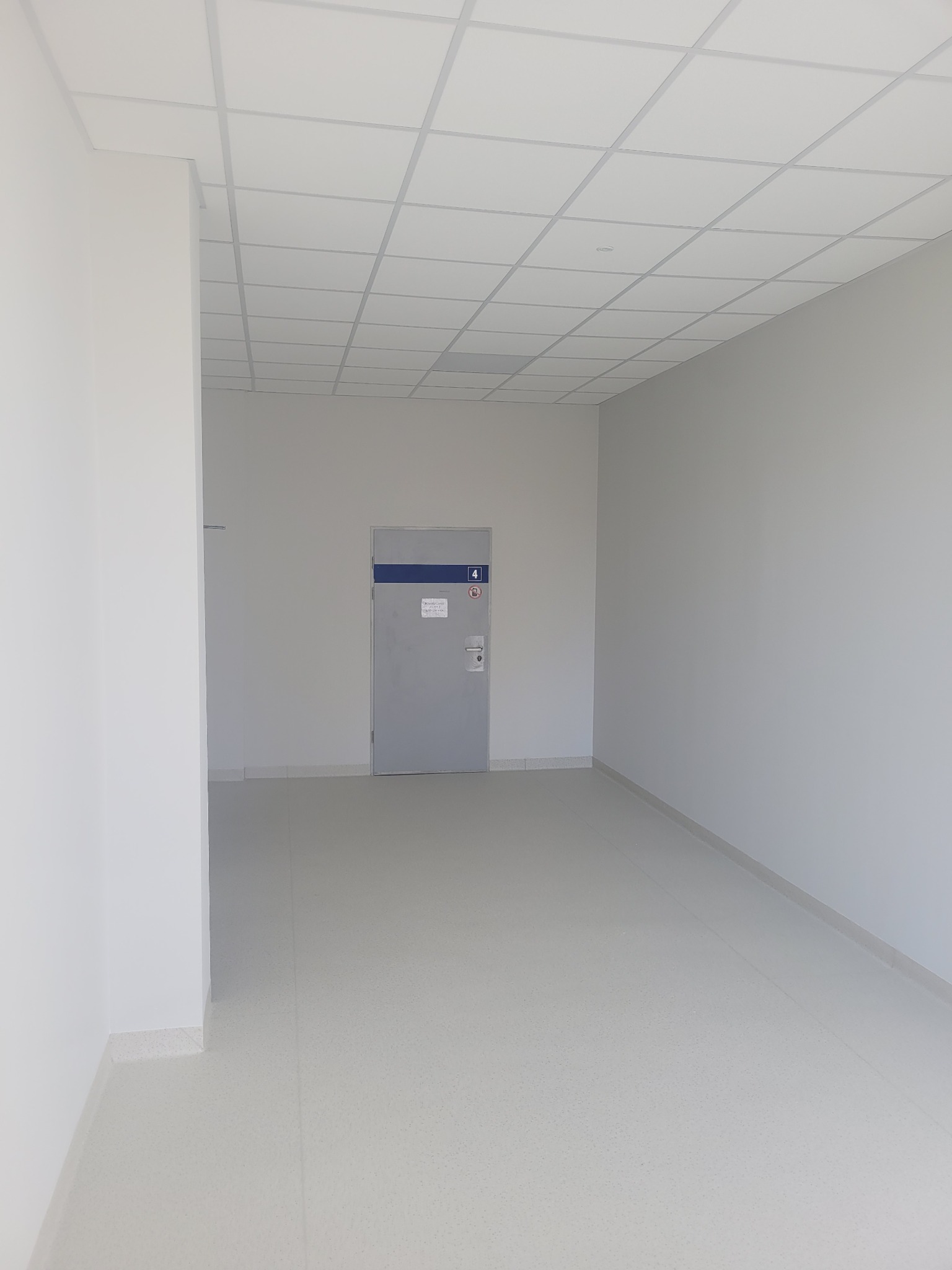 Wnętrze biurowe z jasną podłogą, ścianami i sufitem kasetonowym, szare drzwi z numerem 4 i niebieskim paskiem na górze.