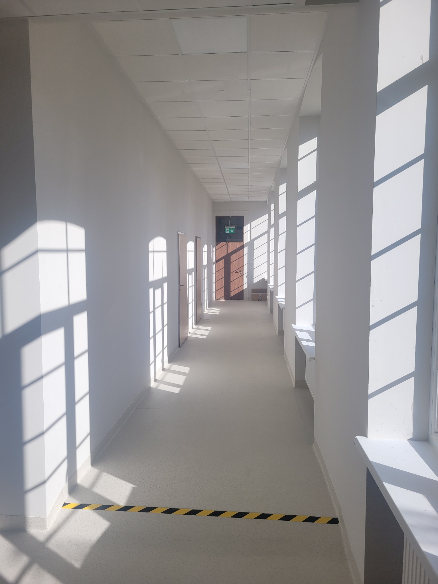 Długi, jasny korytarz biurowy z białymi ścianami i oknami, przez które wpada silne światło słoneczne, tworząc kontrastowe cienie. Na podłodze widoczna taśma ostrzegawcza w czarno-żółte pasy.