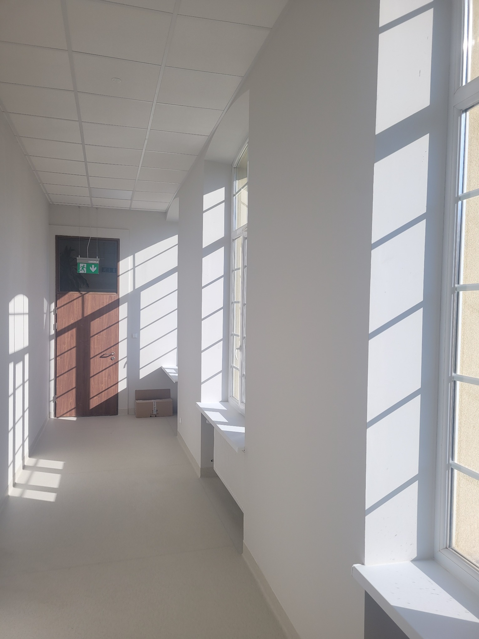 Jasny korytarz biurowy po remoncie z widocznymi promieniami słońca padającymi przez okna, świeżo pomalowane ściany i sufit kasetonowy, brązowe drzwi ewakuacyjne w oddali.
