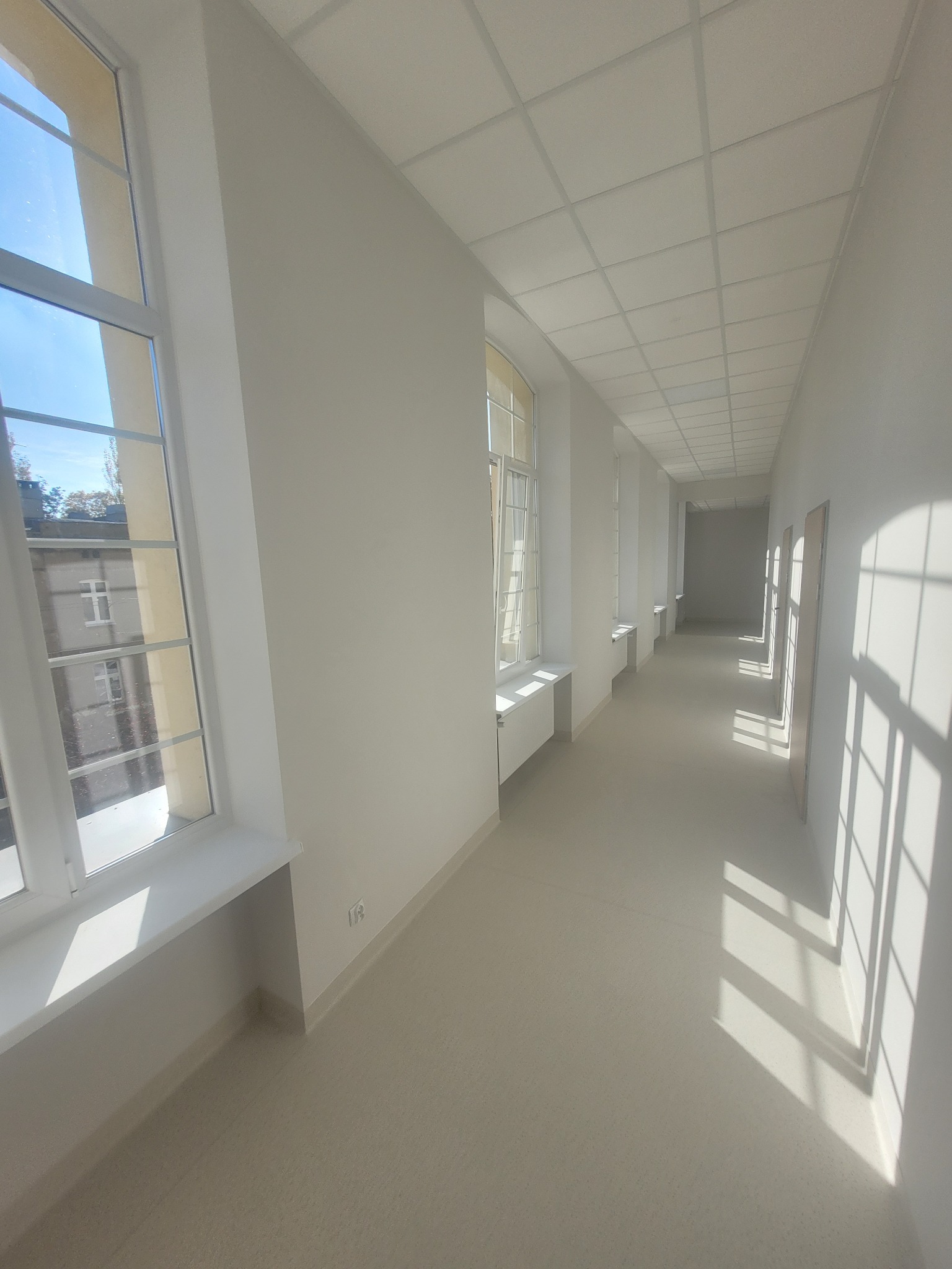 Jasny, długi korytarz biurowy po remoncie, z rzędami okien po lewej stronie i świeżo pomalowanymi ścianami na biało.