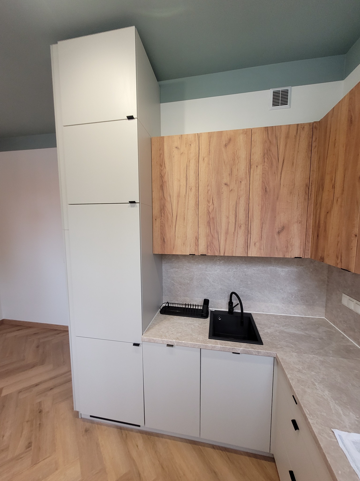 Nowoczesna kuchnia z jasnymi szafkami dolnymi i wysokim słupkiem, drewnopodobne szafki górne, czarny zlew i bateria, marmurkowy blat, parkiet w jodełkę na podłodze, sufit pomalowany na dwa kolory.