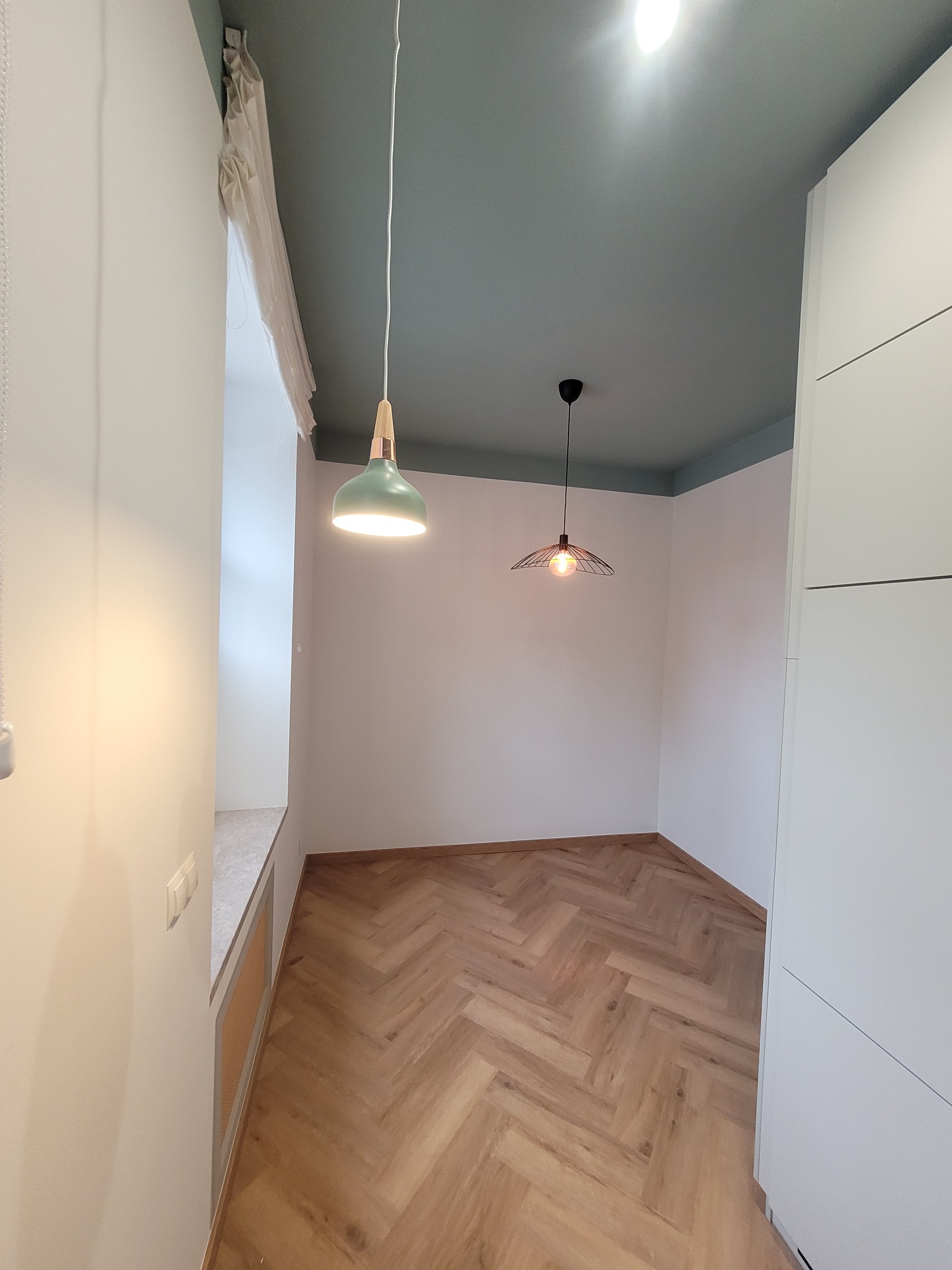 Wnętrze pokoju z parkietem w jodełkę, pomalowanym na zielono sufitem z dwoma wiszącymi lampami o różnych stylach, białymi ścianami i zabudową.