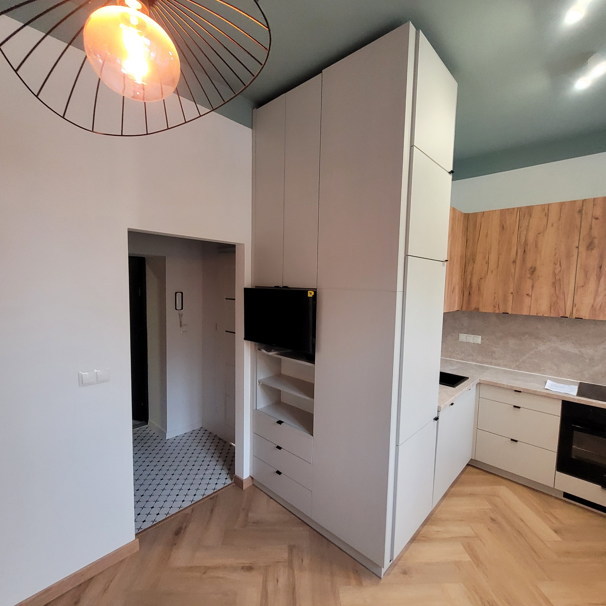 Widok na nowoczesną kuchnię z zabudową meblową w odcieniach bieli i drewna, z telewizorem wkomponowanym w szafę i parkietem ułożonym w jodełkę.