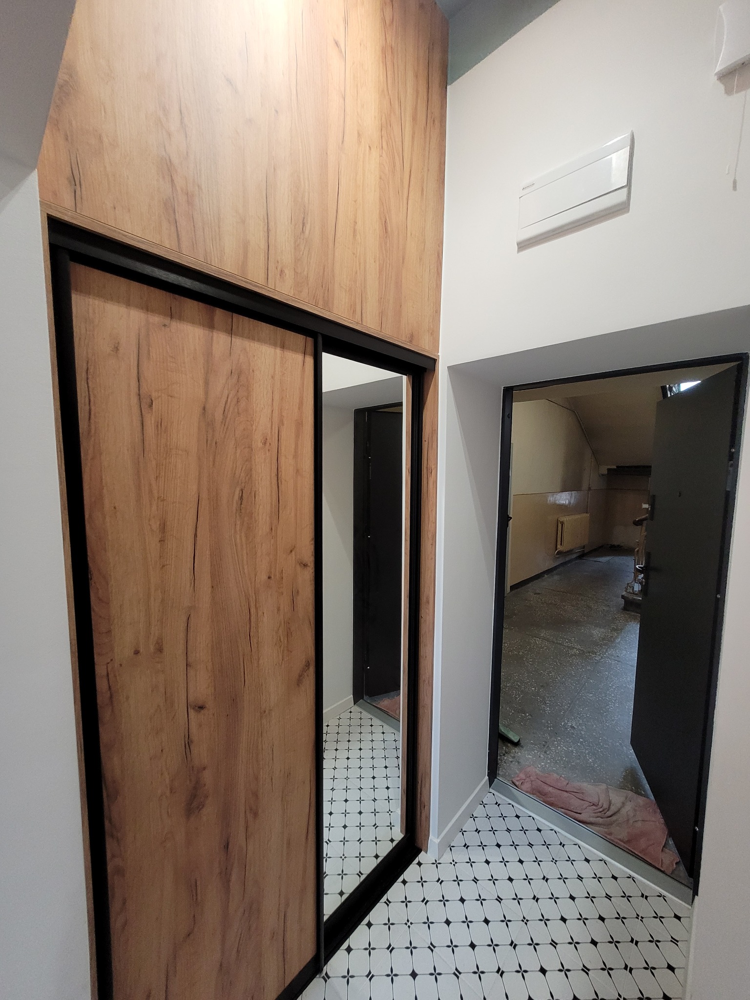 Nowoczesna szafa wnękowa z przesuwnymi drzwiami imitującymi drewno, czarne obramowanie, białe ściany, podłoga w czarno-białą szachownicę, widok na korytarz z otwartymi drzwiami.