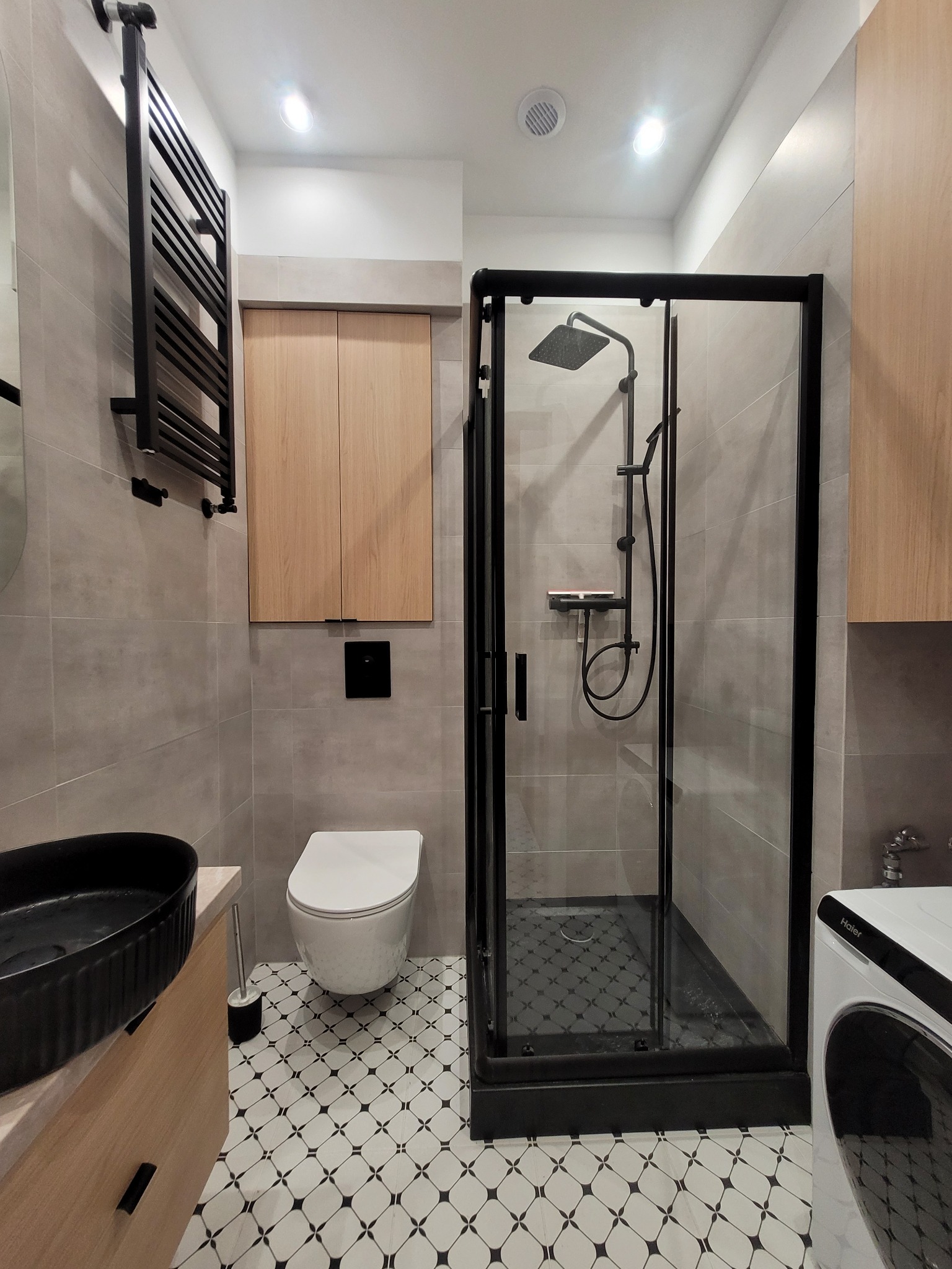 Nowoczesna łazienka z czarną armaturą: kabina prysznicowa typu walk-in z deszczownicą, umywalka nablatowa na szafce z jasnego drewna, grzejnik drabinkowy i biała toaleta w stylu minimalistycznym...