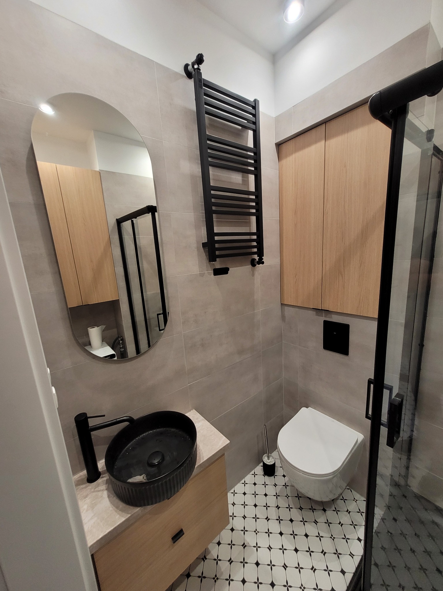 Nowoczesna łazienka z owalnym lustrem, czarną umywalką nablatową, grzejnikiem drabinkowym, szafkami wiszącymi z jasnego drewna i toaletą podwieszaną na tle szarej płytek ściennych i podłogi...