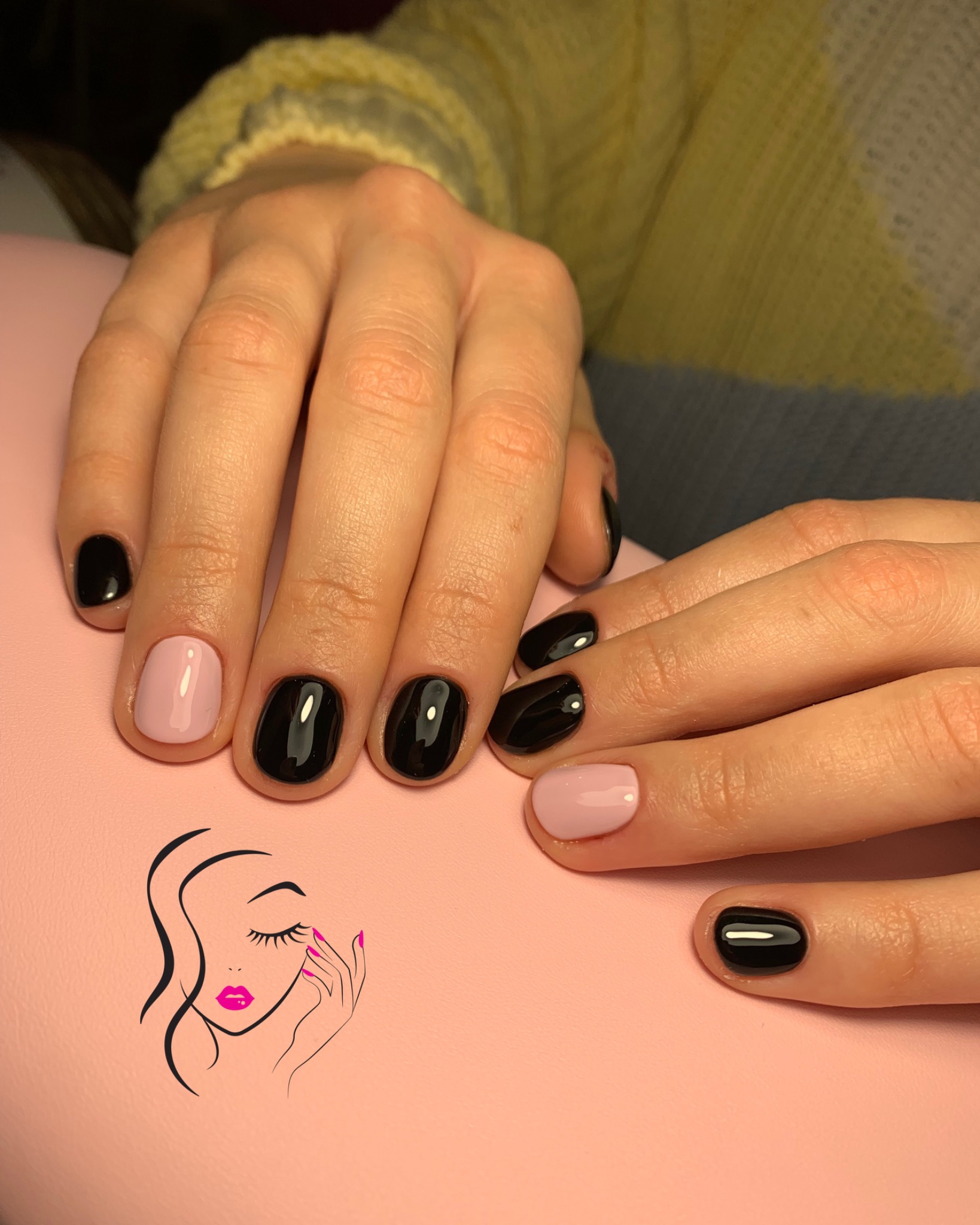 Dłonie z manicure hybrydowym: czarne paznokcie z jednym jasnoróżowym akcentem na każdej dłoni, ułożone na różowym podłożu z nadrukowanym logo salonu kosmetycznego.