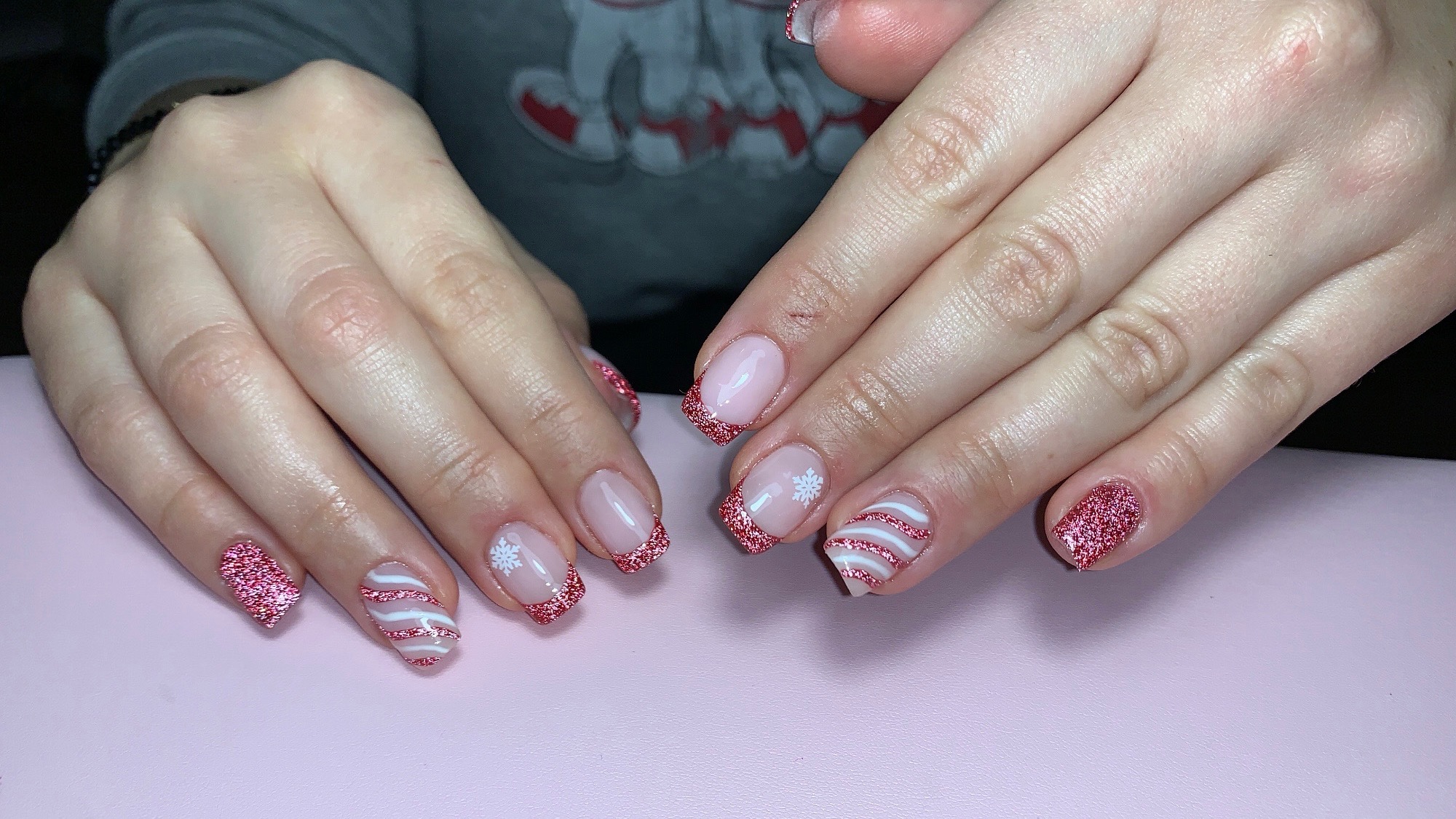 Dłonie z manicure hybrydowym: różowy brokat na końcówkach, białe paski i śnieżynki na jasnoróżowym tle.