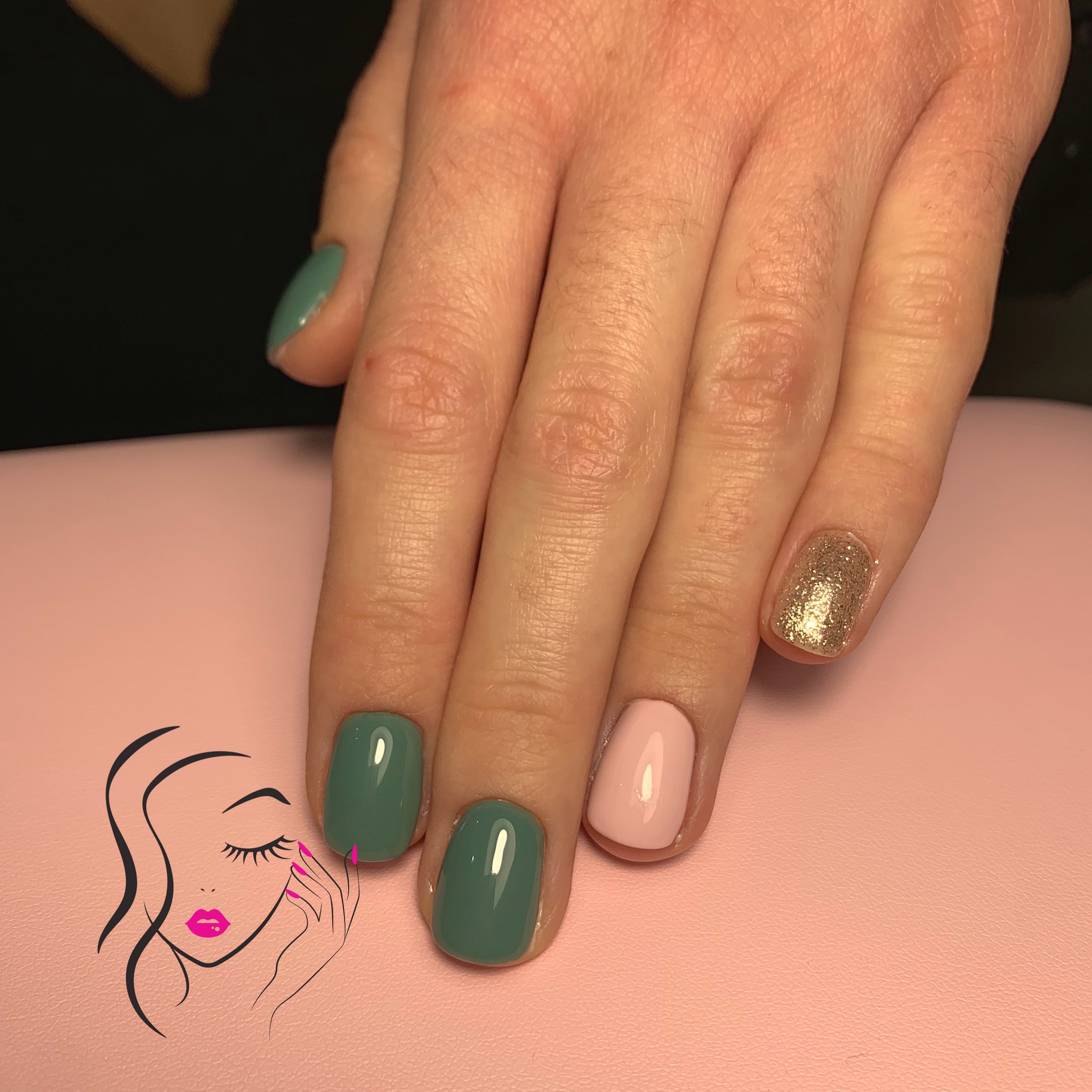 Krótkie paznokcie pomalowane lakierami w kolorach: butelkowa zieleń, pudrowy róż i brokatowe złoto, na tle różowej podkładki z nadrukowanym logo salonu.