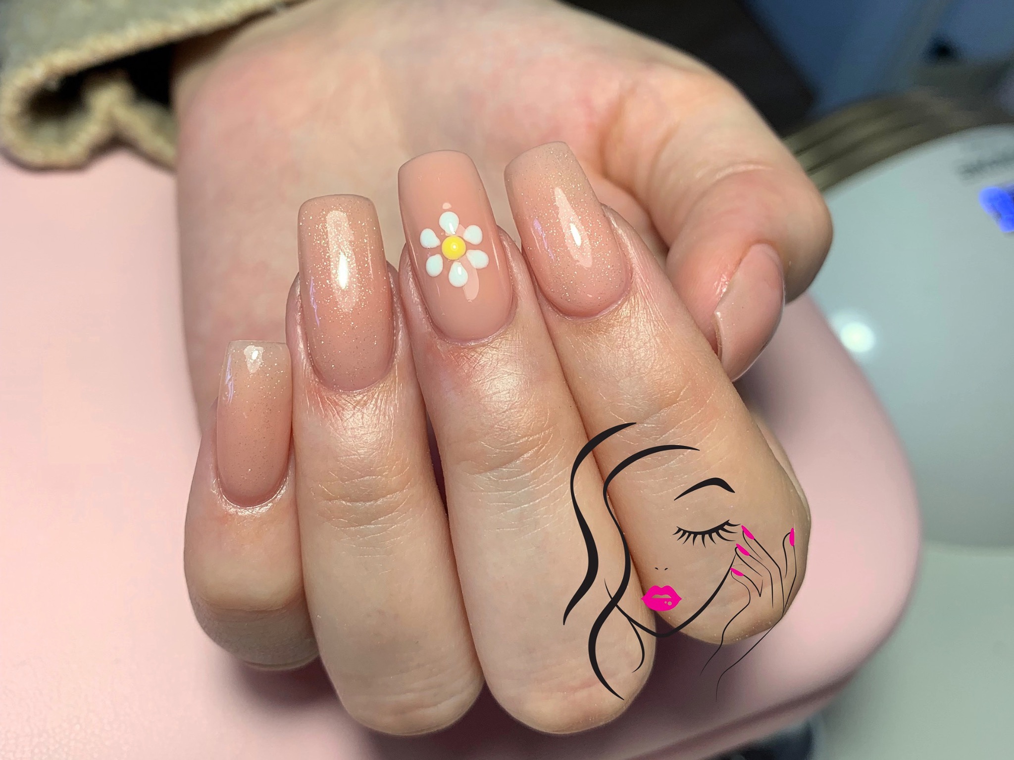 Dłoń z przedłużonymi, kwadratowymi paznokciami w odcieniach nude z brokatem, jeden paznokieć ozdobiony biało-żółtym kwiatkiem, na tle różowy blat i grafika kobiety z różowymi ustami.