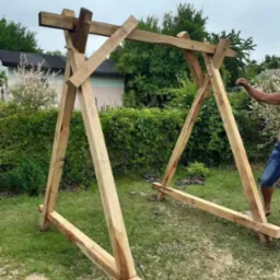 Drewniana konstrukcja w ogrodzie, prawdopodobnie altana lub pergola w trakcie budowy, widoczne świeże wióry, tło z zielenią i domem.