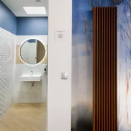 Nowoczesna toaleta z okrągłym lustrem z podświetleniem LED, umywalką nablatową, drewnianą podłogą i grzejnikiem dekoracyjnym na tle abstrakcyjnej tapety w odcieniach niebieskiego.