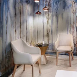 Jasne krzesła z drewnianymi nogami na tle ściany z motywem leśnym, oświetlone trzema miedzianymi lampami wiszącymi, stolik kawowy z ciemnym blatem.