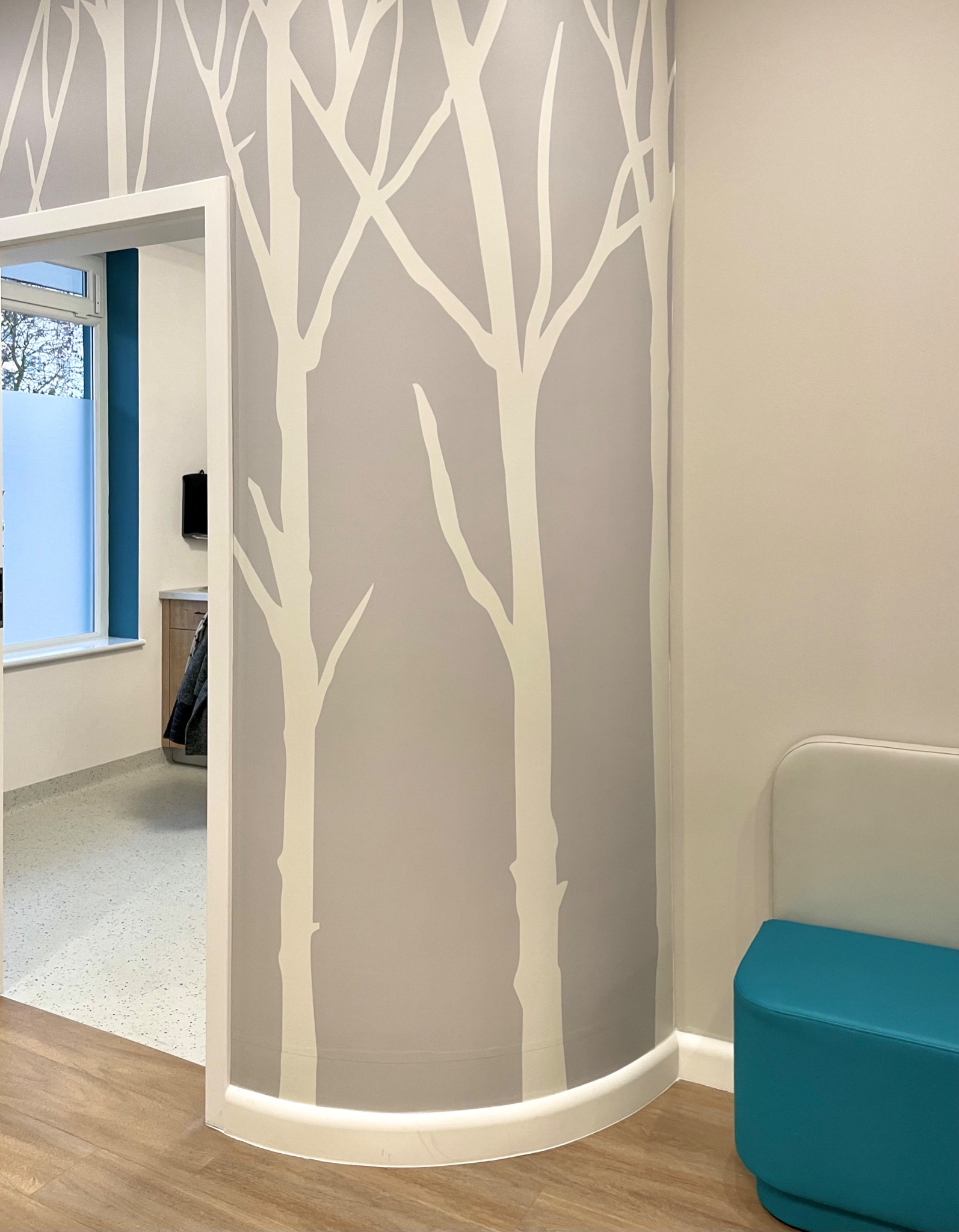Korytarz w biurze z szarą ścianą zdobioną białymi, stylizowanymi drzewami, widok na sąsiednie pomieszczenie przez białe drzwi, turkusowy puf przy ścianie.