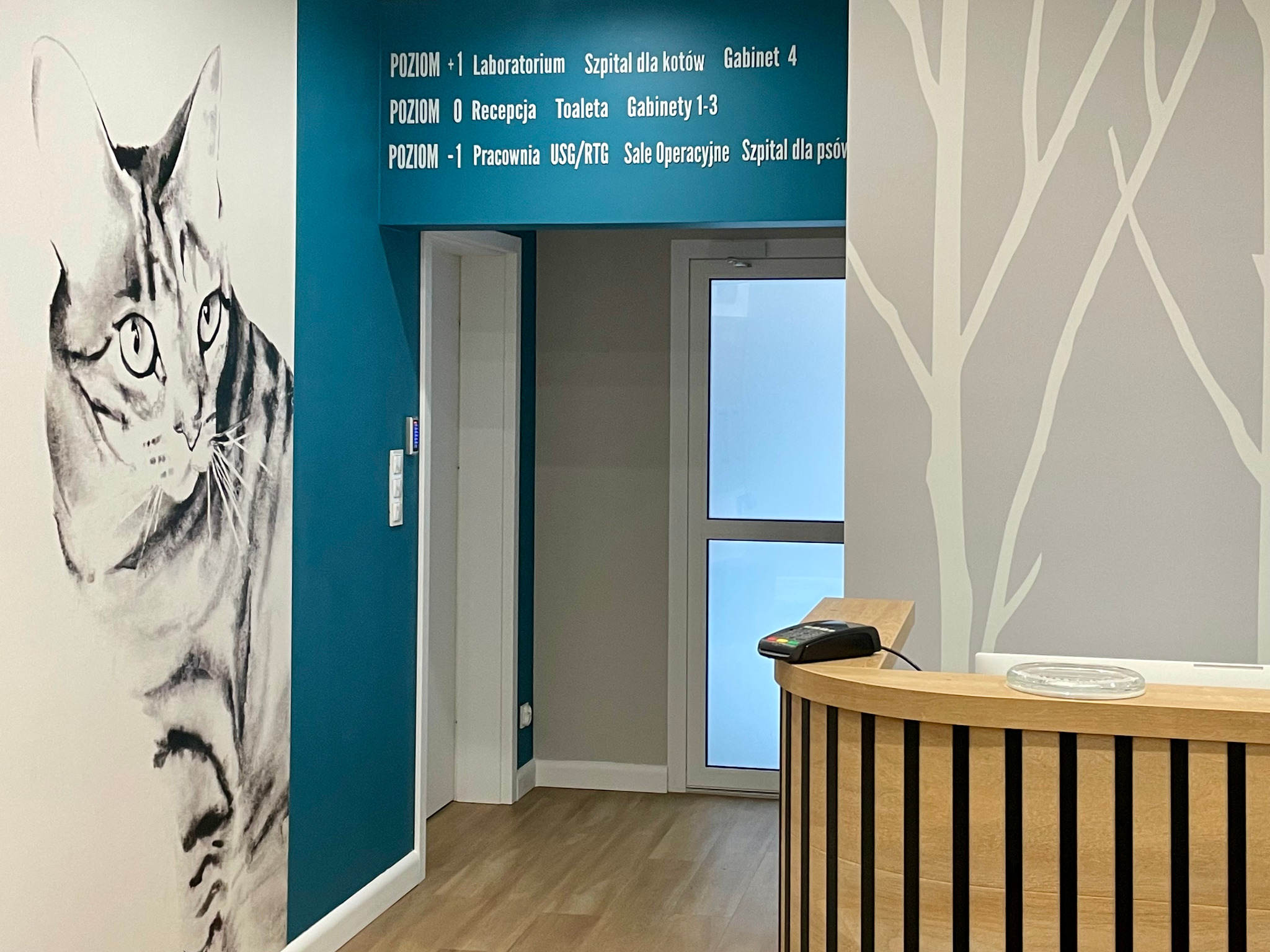 Recepcja w klinice weterynaryjnej z grafiką kota na ścianie, drewniana lada z terminalem płatniczym i tablicą informacyjną o poziomach i gabinetach.
