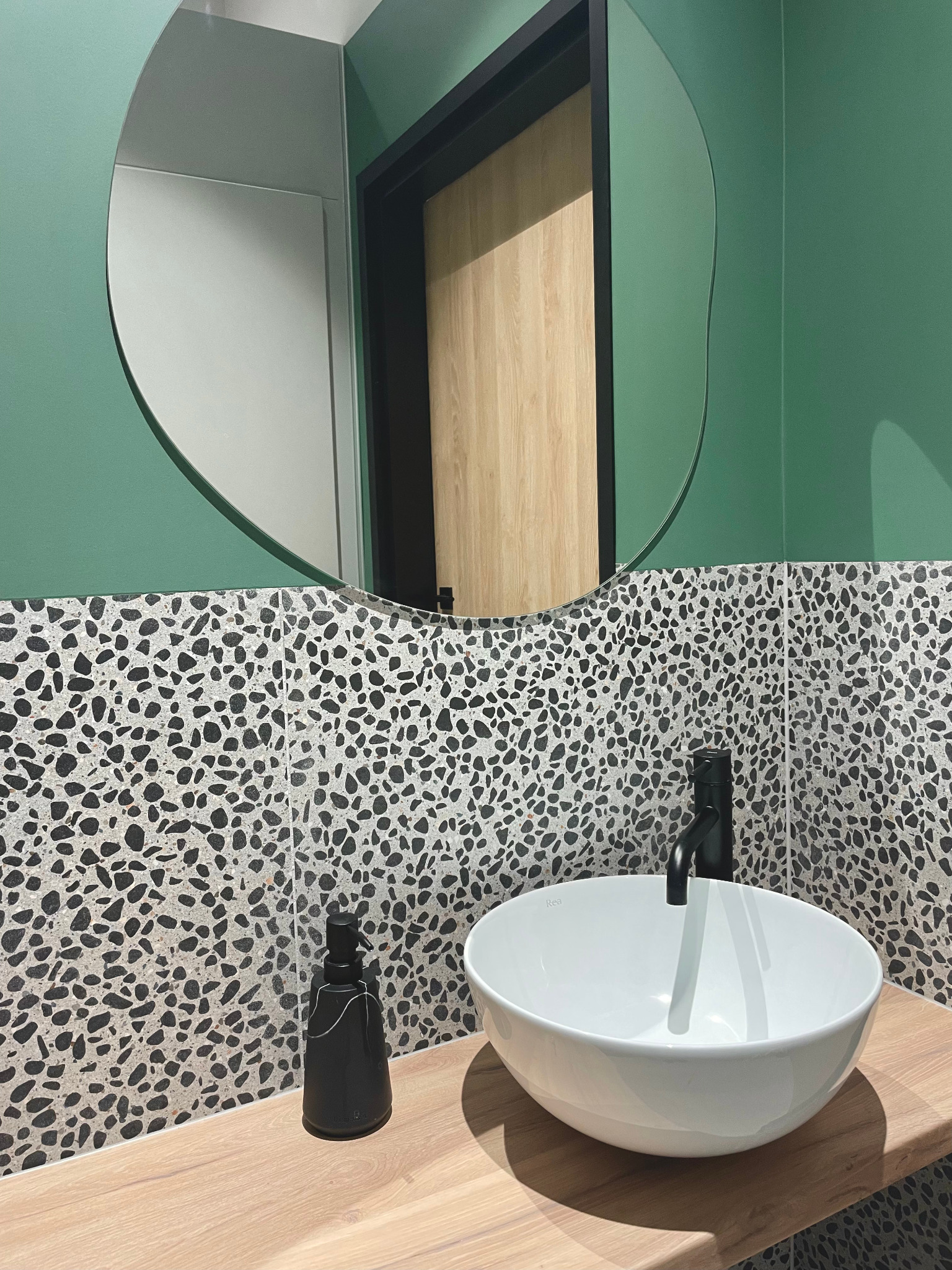 Nowoczesna łazienka z zielonymi ścianami, umywalką nablatową i lustrem o nieregularnym kształcie. Na blacie stoi czarny dozownik do mydła.