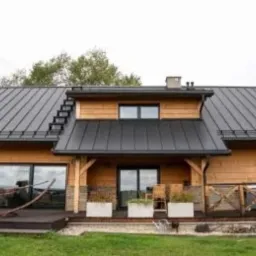 Drewniany dom jednorodzinny z dużymi przeszkleniami, tarasem z hamakiem i metalowym dachem w kolorze antracytowym. Widoczne elementy konstrukcyjne wykonane z drewna. Opis 'POLSKA' sugeruje...