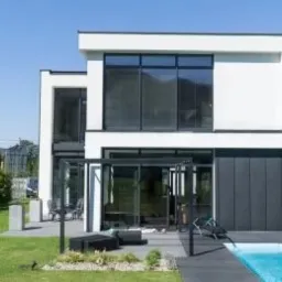 Nowoczesny, dwukondygnacyjny dom z płaskim dachem, dużymi przeszkleniami w czarnych ramach, z widocznym basenem i zadbanym trawnikiem w słoneczny dzień.