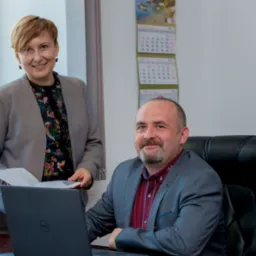 Dwie osoby w biurze: kobieta w kwiecistej bluzce i szarej marynarce trzyma dokumenty, mężczyzna w szarej marynarce siedzi przy laptopie na czarnym skórzanym fotelu, za nimi kalendarz i geometryczna...