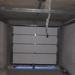 Otwarta segmentowa brama garażowa w kolorze białym, widoczne prowadnice i sprężyny, surowy betonowy garaż w trakcie wykańczania.