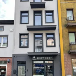 Elewacja budynku z szarymi oknami PCV i napisem SHOWROOM nad wejściem, widoczne detale architektoniczne i sąsiednia kamienica z ż&oacute;łtym akcentem.