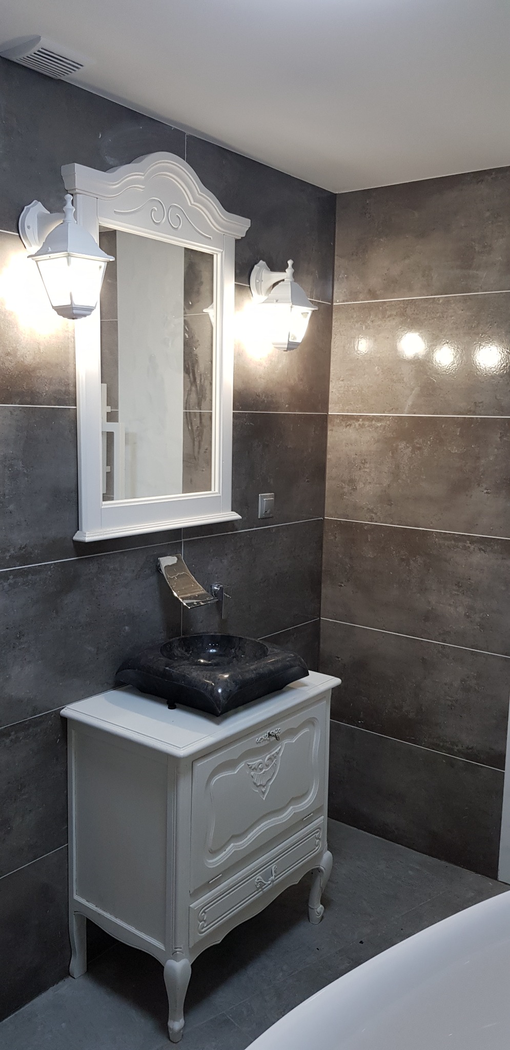 Wykończona łazienka z szarą płytką imitującą kamień, białą szafką pod umywalkę w stylu retro z czarną, kwadratową umywalką nablatową i srebrną baterią kaskadową, lustro w białej ramie z kinkietami...