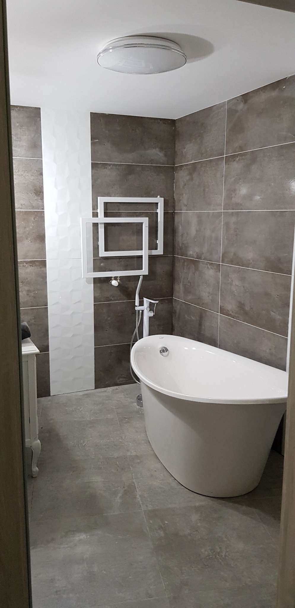 Nowoczesna łazienka w Gdańsku z wolnostojącą białą wanną, szarobrązowymi płytkami, dekoracyjną białą ścianą i grzejnikiem drabinkowym w kształcie kwadratów.