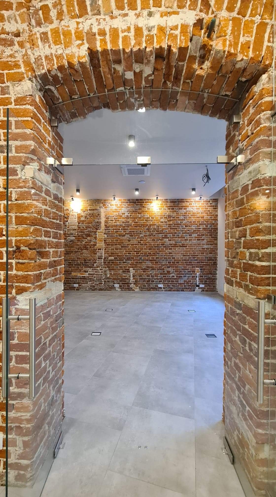 Wnętrze odnowionego pomieszczenia z ceglanymi ścianami, łukowym przejściem i szarą podłogą z płytkami. Widoczne szklane drzwi z metalowymi uchwytami.