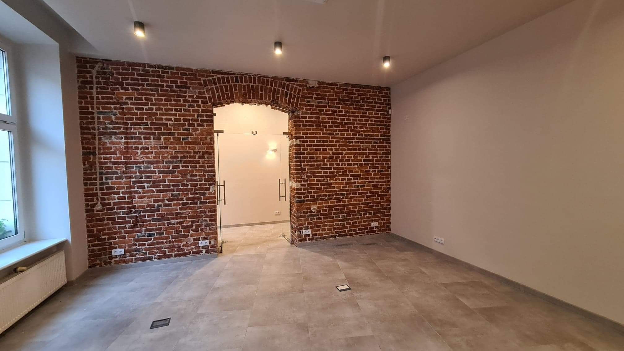 Wykończone wnętrze biurowe z ceglaną ścianą z łukowym przejściem oszklonymi drzwiami, szara podłoga z płytek, białe ściany i sufit z trzema punktowymi lampami.