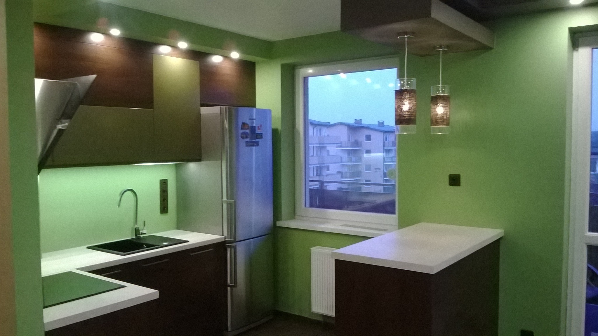 Nowoczesna kuchnia w odcieniach zieleni i brązu z białymi blatami, oświetlona lampami wiszącymi, z widokiem na budynki mieszkalne za oknem.