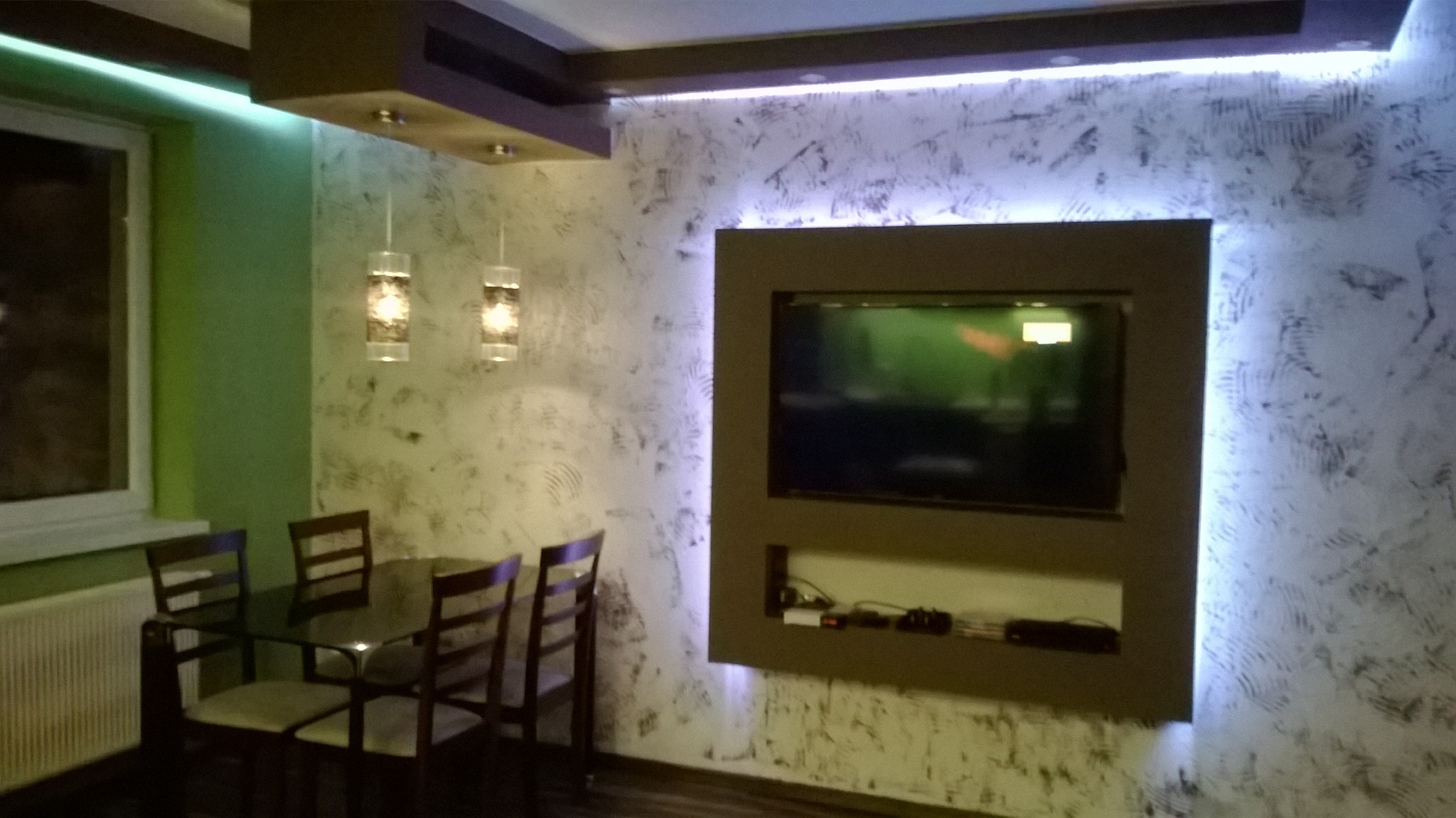 Nowoczesne wnętrze pokoju z dekoracyjną ścianą, podświetlanym sufitem i telewizorem w obudowie, stół z krzesłami w tle okna.