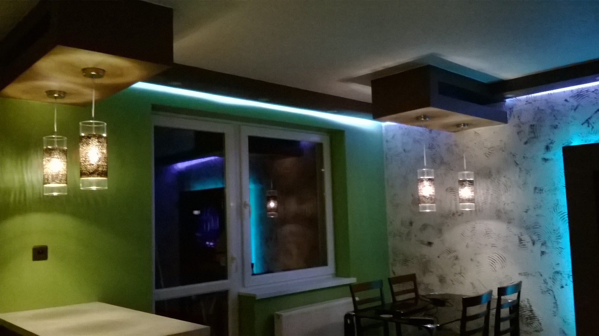 Nowoczesna kuchnia z oknem, oświetleniem LED i dekoracyjną ścianą, uzupełniona wiszącymi lampami o cylindrycznym kształcie.