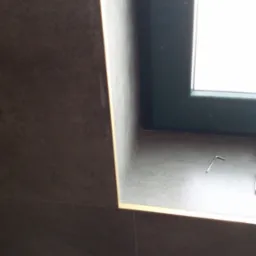 Fragment ściany z ciemnymi płytkami obok okna z szarą ramą, widoczny profil wykończeniowy w kolorze złota i klucz imbusowy leżący na parapecie.