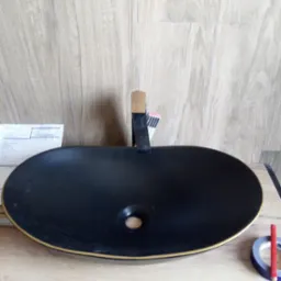 Czarna, owalna umywalka nablatowa z złotym rantem, zamontowana bateria z drewnianym elementem, widoczny ołówek i taśma klejąca na blacie imitującym drewno, tło stanowią panele ścienne.