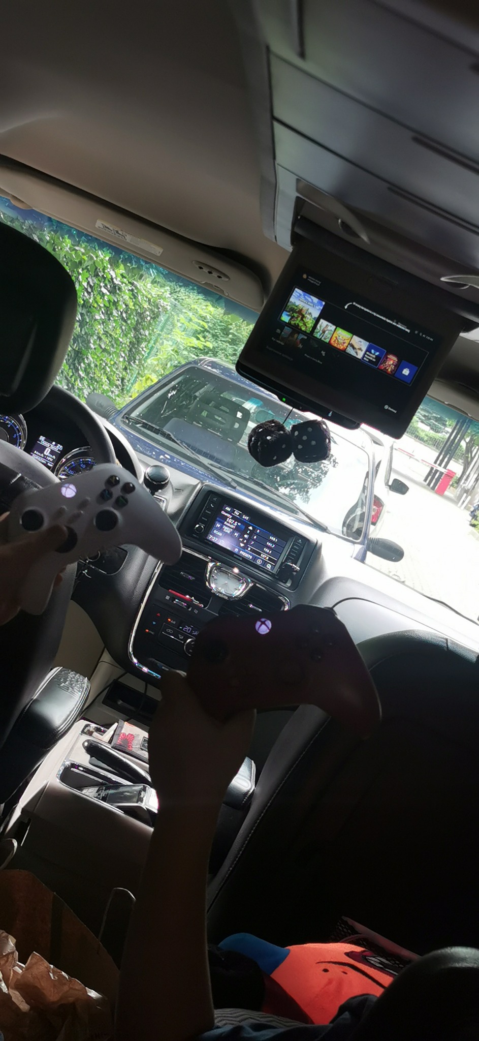 Wnętrze minivana z wbudowanym ekranem i dwoma kontrolerami Xbox w rękach pasażerów na tylnych siedzeniach, widoczne kostki do gry wiszące na lusterku wstecznym.