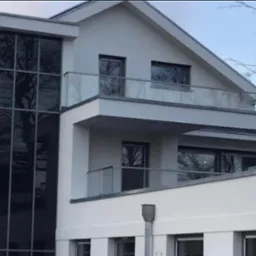 Nowoczesny dom z dużymi przeszkleniami i balkonem z szklaną balustradą, widoczne odbicia drzew w oknach.