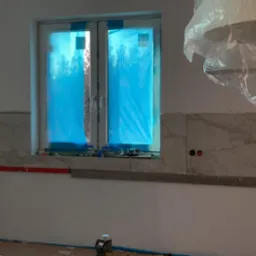Układanie płytek imitujących marmur na ścianie w kuchni, widoczne zabezpieczone okno folią, poziomica laserowa na podłodze i fragment instalacji elektrycznej.
