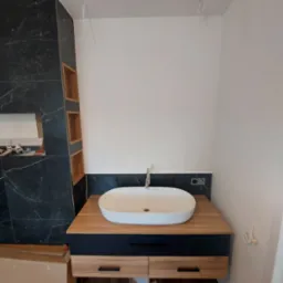 Nowoczesna biała umywalka nablatowa na drewnianej szafce z czarnymi frontami szuflad, marmurowe płytki na ścianie z wnękami, widoczne kable elektryczne.