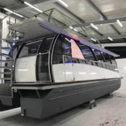 Nowoczesny katamaran z przeszkloną kabiną i spiralnymi schodami ze stali nierdzewnej, oświetlony niebieskimi diodami LED, na kółkach w hali produkcyjnej.