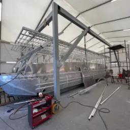 Wnętrze hali produkcyjnej z konstrukcją aluminiową łodzi katamaranu w trakcie budowy, widoczny wózek narzędziowy Sherman, suwnica i rusztowanie.