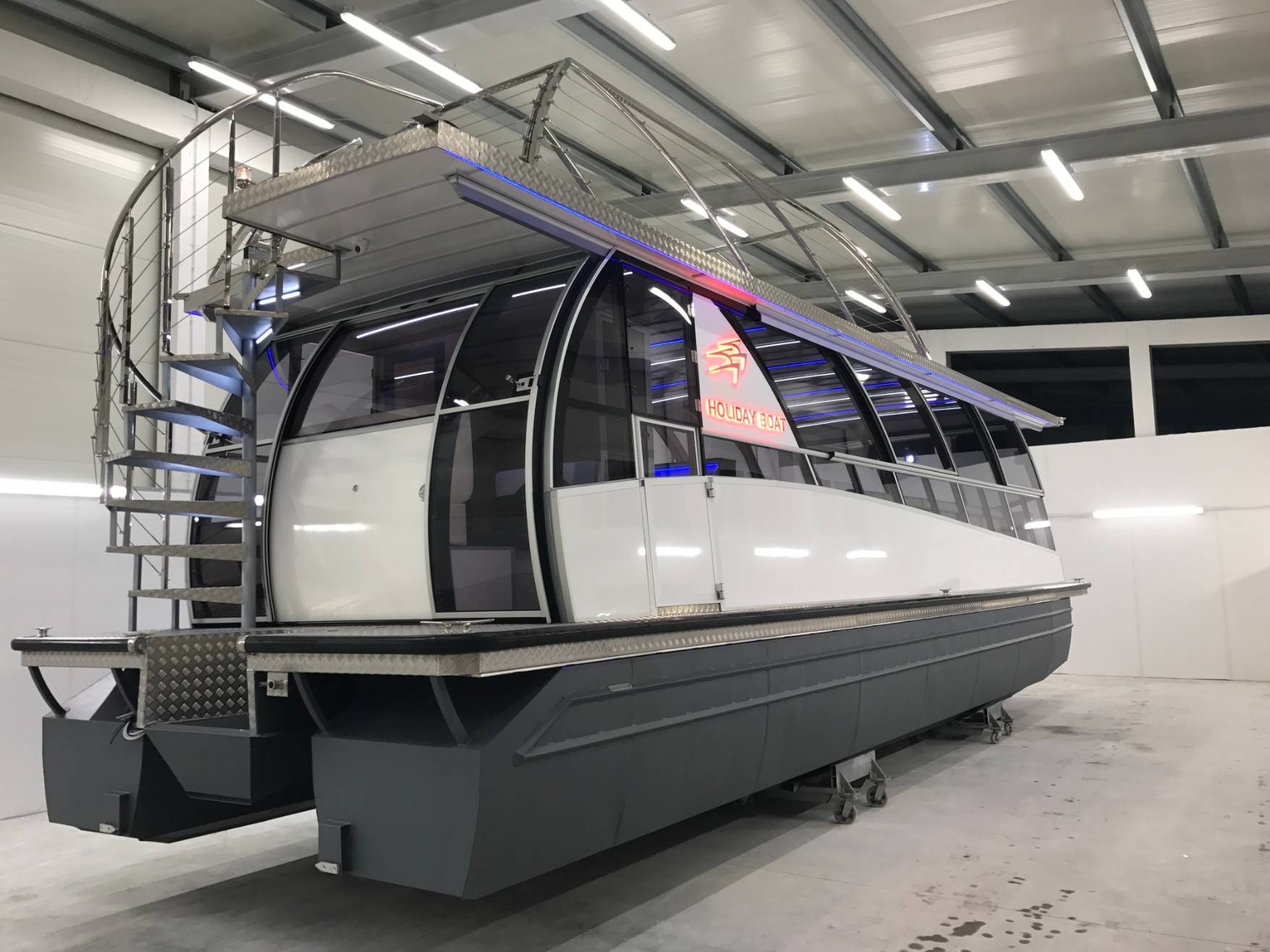 Nowoczesny katamaran z przeszkloną kabiną i spiralnymi schodami ze stali nierdzewnej, oświetlony niebieskimi diodami LED, na kółkach w hali produkcyjnej.