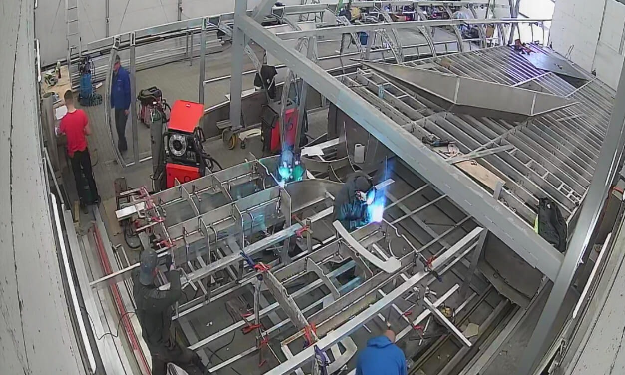 Spawanie metalowej konstrukcji szkieletowej w hali produkcyjnej, widoczni spawacze w maskach i kombinezonach, iskry, czerwona spawarka, inni pracownicy w tle.