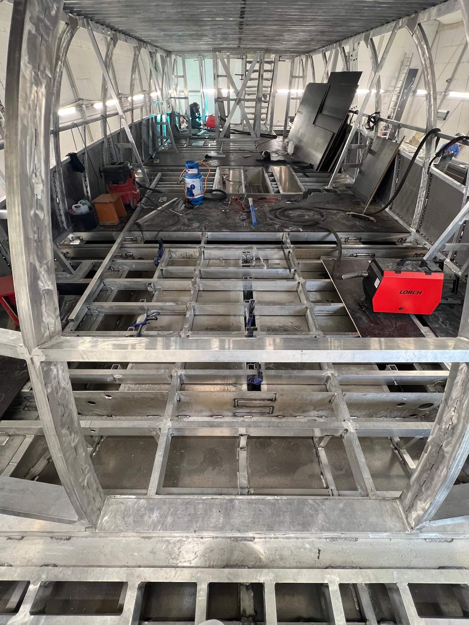 Wnętrze metalowej konstrukcji w trakcie budowy, widoczne spawane elementy szkieletu, sprzęt spawalniczy i akcesoria, w tle pracownik przy drabinie.