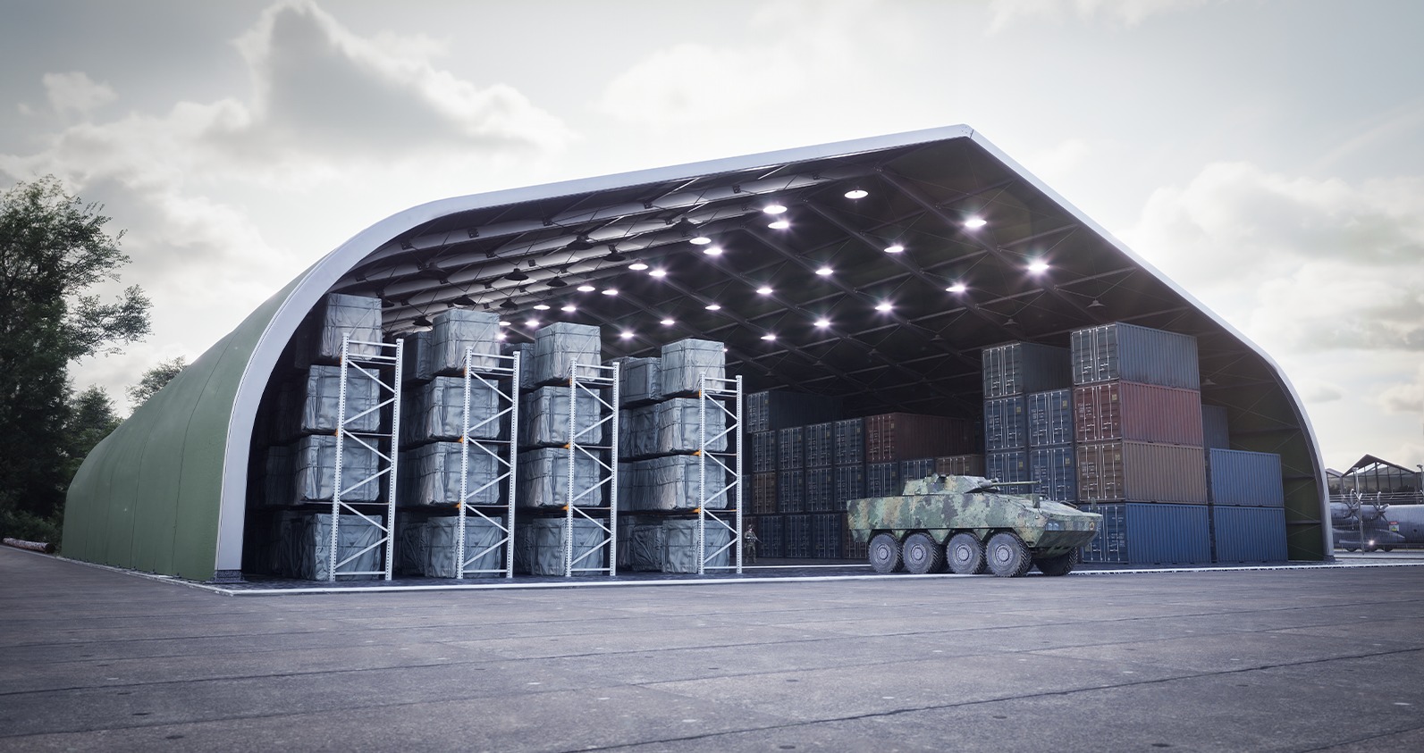 Duży namiotowy magazyn z kontenerami i pojazdem opancerzonym wewnątrz, oświetlony licznymi lampami sufitowymi.