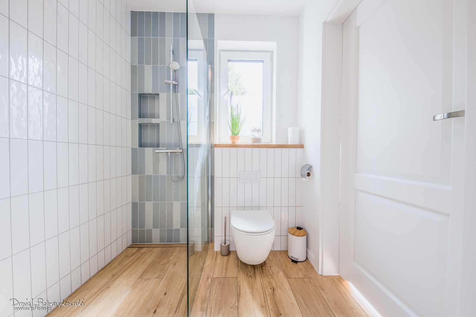 Jasna łazienka z kabiną prysznicową, drewnianą podłogą i białymi płytkami na ścianach, akcent szarych płytek w strefie prysznica, okno z rośliną na parapecie.