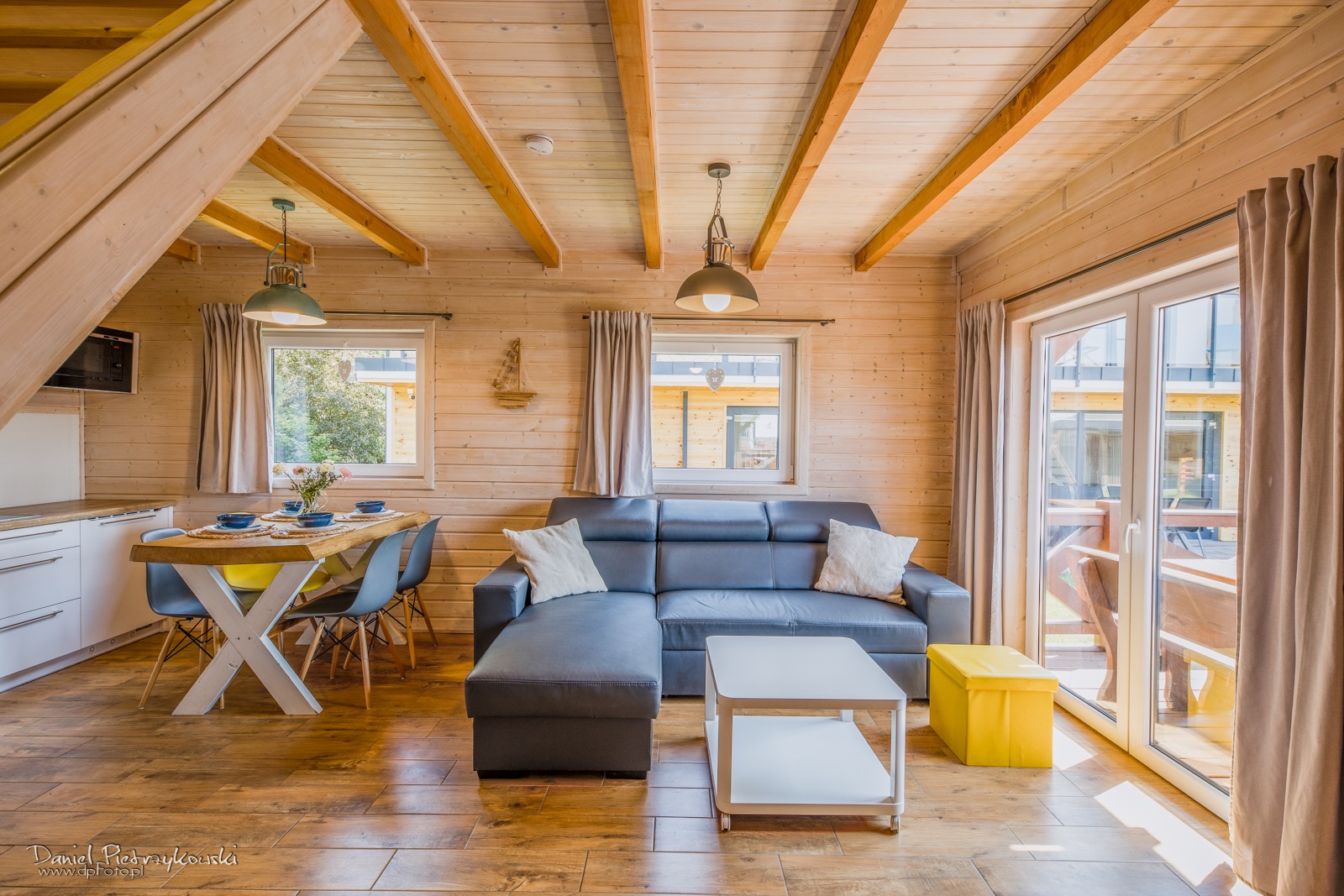 Jasne, drewniane wnętrze domku letniskowego z szarą sofą narożną, białym stolikiem kawowym, stołem z kwiatami i krzesłami, oraz żółtym pufem, oświetlone wiszącymi lampami.