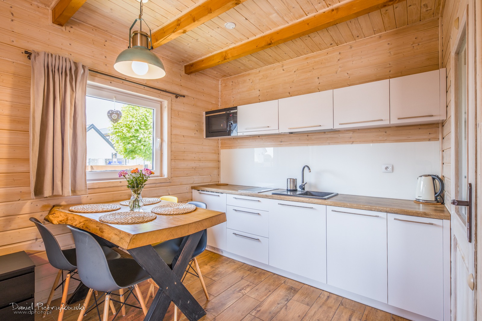 Jasna, drewniana kuchnia z białymi szafkami, drewnianym blatem i stołem z blatem z surowego drewna. W oknie wazon z kwiatami i dekoracja w kształcie serca.