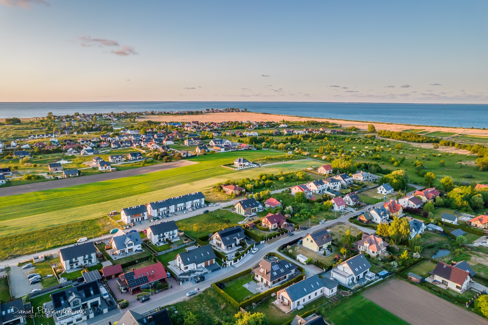 Panoramiczny widok z lotu ptaka na nadmorską miejscowość z domami jednorodzinnymi, polami uprawnymi i linią brzegową w tle, uchwycony w ciepłym świetle wczesnego poranka.