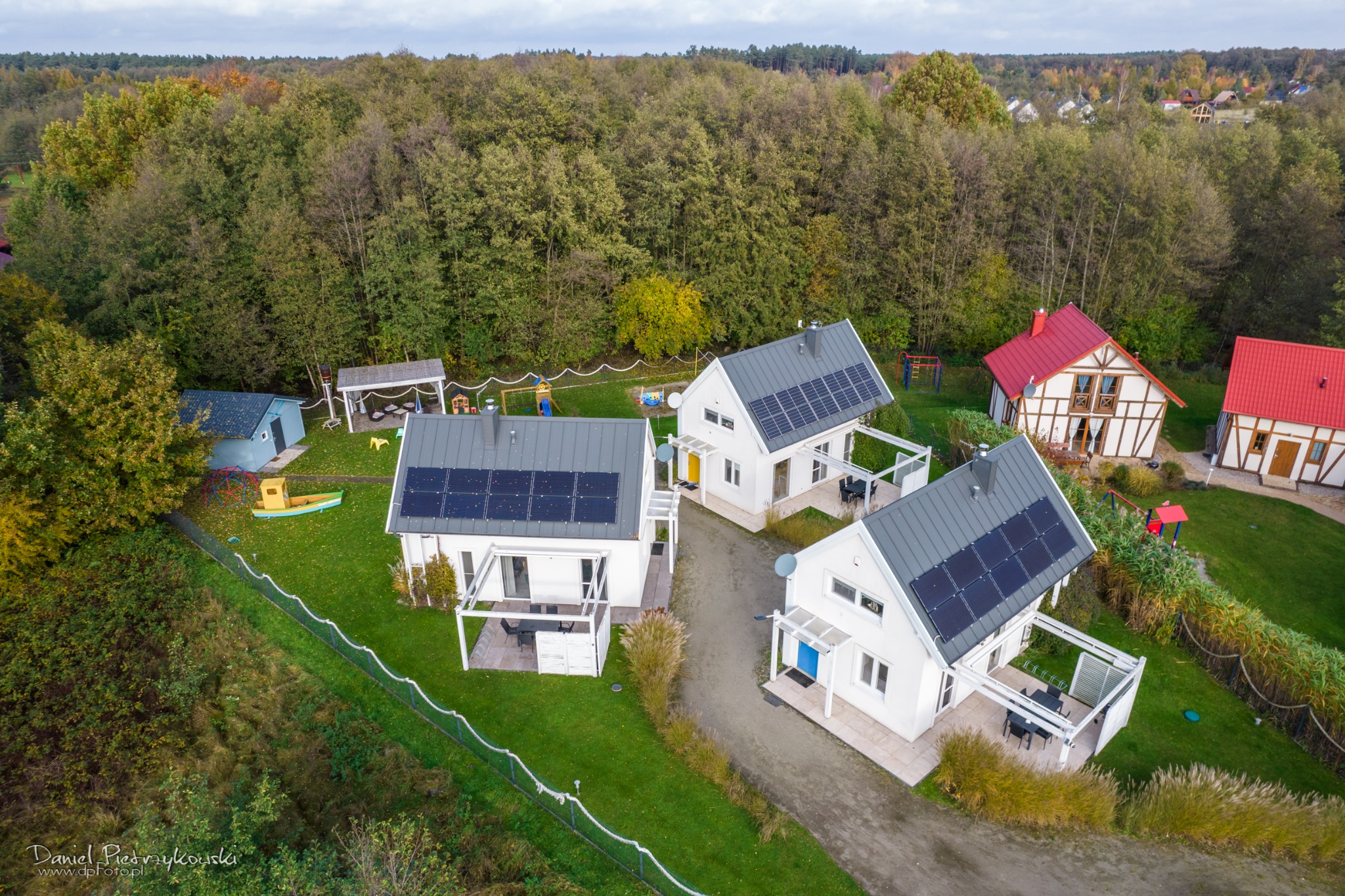 Panoramiczny widok z lotu ptaka na nowoczesne domy z panelami słonecznymi, otoczone zielenią i lasem.