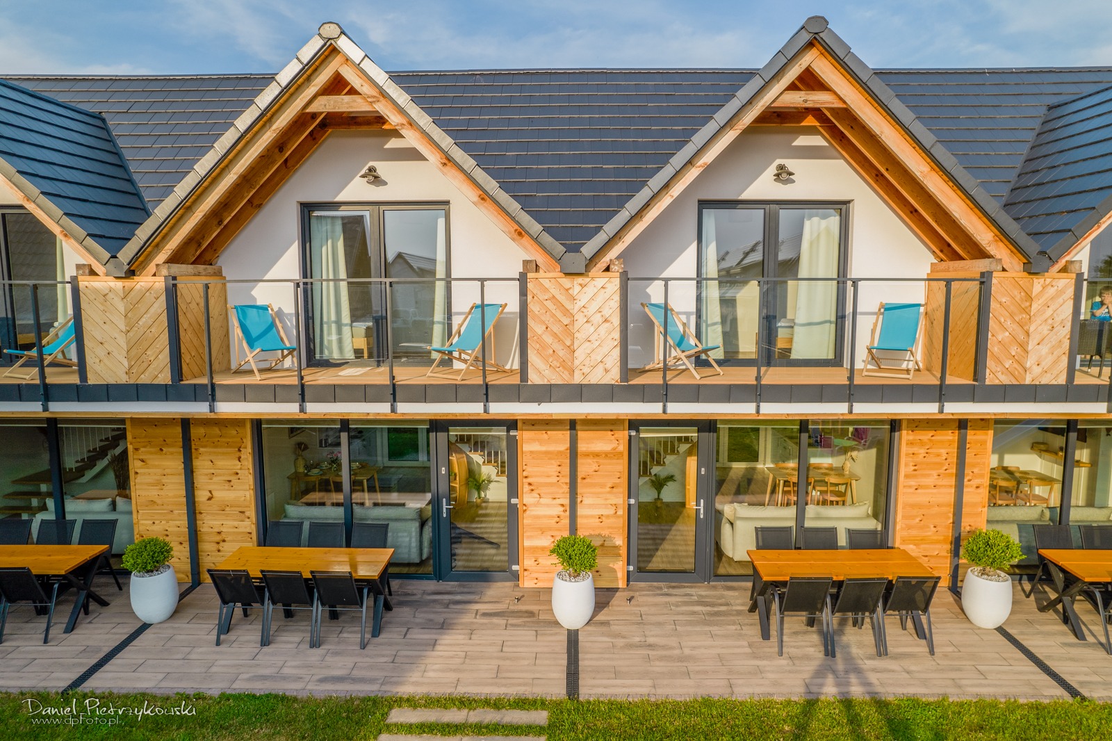Nowoczesny dwupiętrowy budynek z drewnianą elewacją, balkonami z turkusowymi leżakami i tarasem z zestawami stołów i krzeseł, widok z lotu ptaka.